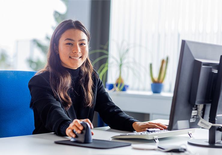 A young woman sits at a desk; there is a computer on it and the woman has one hand on the mouse and one hand on the keyboard.