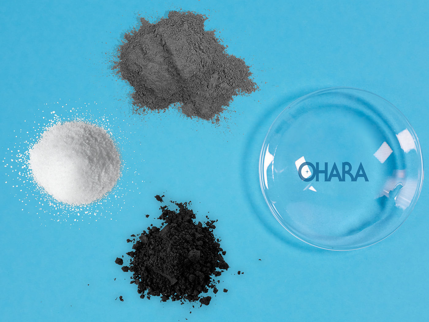 Das Bild hat einen hellblauen Hintergrund, auf diesem sind ein weißes, ein graues und ein schwarzes Pulverhäufchen sowie ein Glasschälchen zu sehen. Auf dem Glasschälchen steht OHARA in blauen Buchstaben.